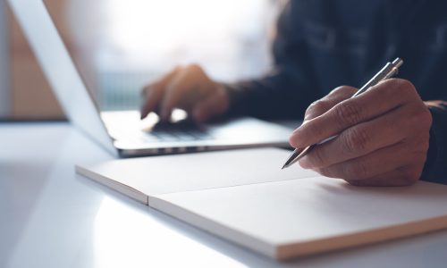 Casual business man, entrepreneur writing on paper notebook, working on laotop computer in modern office, close up. Student studying online course and lecturing on notepad.