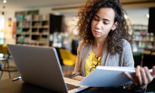Young afro american woman sitting at table with books and laptop for finding information. Young student taking notes from laptop and books for her study in library.