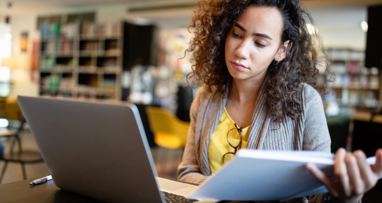 Young afro american woman sitting at table with books and laptop for finding information. Young student taking notes from laptop and books for her study in library.